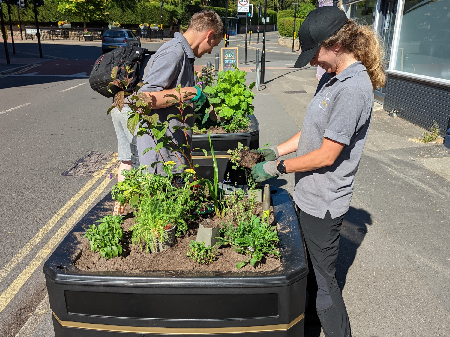 Two people wearing grey uniforms and green gloves are planting herbs and small plants in large black planters positioned on a roadside. The planters contain soil and various green plants, and the individuals are working with gardening tools under bright sunlight. A street with double yellow lines and buildings is visible in the background.