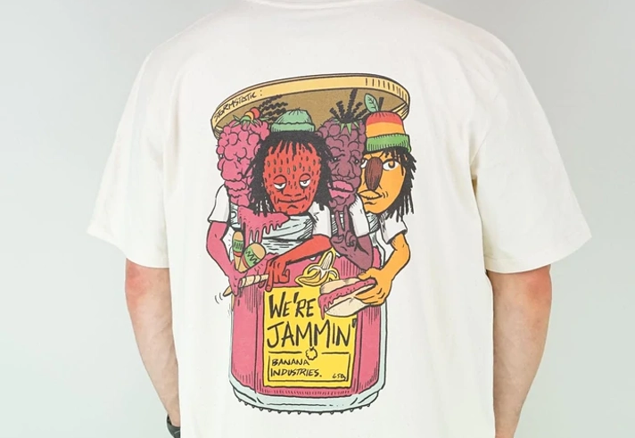 A man stands with his back to the camera, wearing a baseball bat, a white t-shirt with a colourful back print and green trousers. The design on the shirt is four anthropomorphic fruits busting out of a jam jar. They look like members of a band. The yellow label on the jar reads 'We're jammin''.