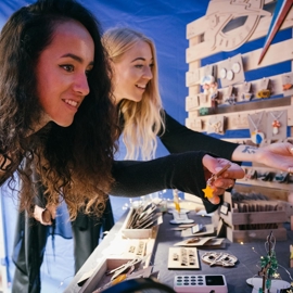 Two people browsing a craft stall displaying handmade wooden items and jewelry. The table is filled with small decorative pieces, and a wooden display rack holds various ornaments and accessories. The setting appears to be under a tent with bright lighting.