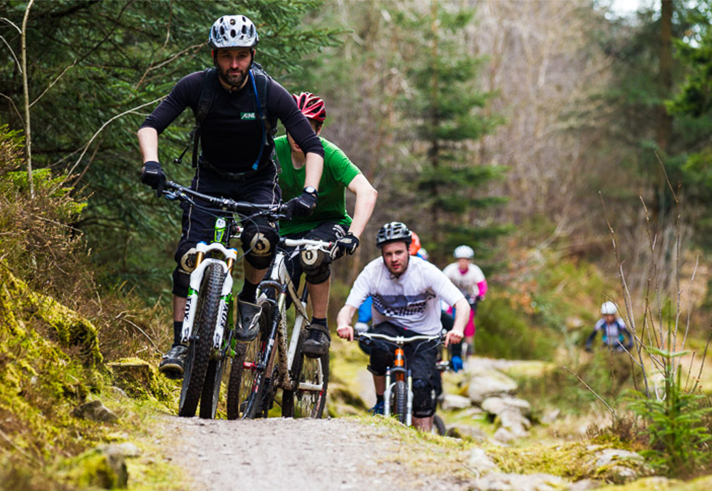 Six or so people, on mountain bikes, ride along a track in a wooded area.