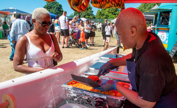 Food stall at an outdoor event with a person serving rice from metal trays under a red canopy decorated with patterned fabric. A crowd of people, including families with children and strollers, is visible in the background on a grassy area with trees and a blue food truck.