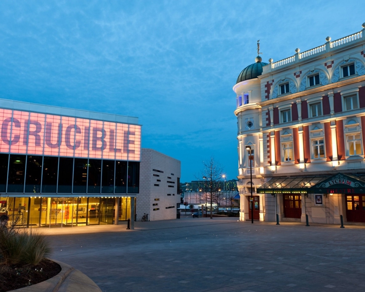 Evening view of Sheffield’s Crucible Theatre and the Lyceum Theatre. The Crucible building has a modern glass façade with large illuminated letters spelling “CRUCIBLE” in pink tones. To the right, the Lyceum Theatre features ornate architecture with columns, decorative details, and warm lighting highlighting its dome and entrance canopy. The sky is a deep blue, and the open plaza in front is quiet and spacious.
