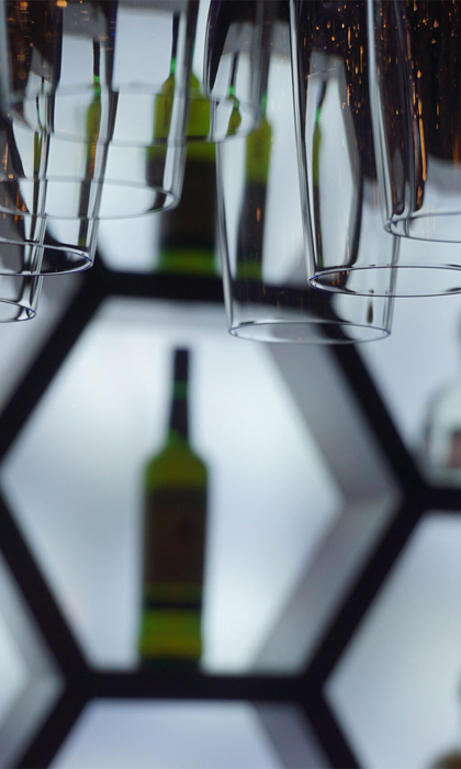 Close-up of an illuminated bar display featuring hexagonal shelves with bottles of liquor arranged inside. Several upside-down hanging glasses are in the foreground, creating a layered effect. The shelves have a bright white backlight, contrasting with the dark surroundings and highlighting the shapes of the bottles and glassware.