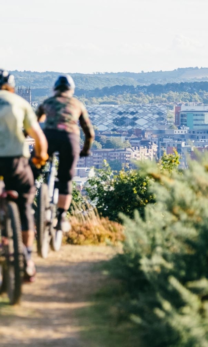 Two people are mountain biking along a dirt track that is flanked with heather on both sides. In the distance the Sheffield skyline is visible.