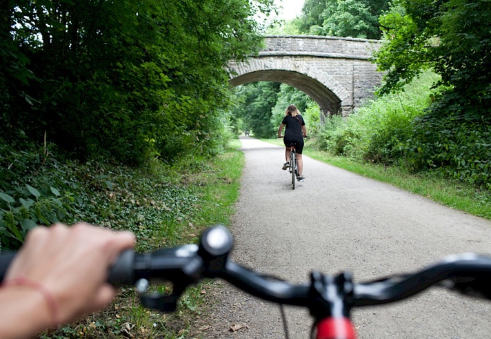 A hand holding the handlebars on their bike, with a cyclist riding along the Monsal Trail in front.