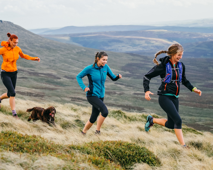 Three people running along a grassy hillside in an open moorland landscape, accompanied by a brown dog. The runners are wearing outdoor athletic clothing, including jackets and leggings, and the terrain is covered with tufts of grass and low vegetation. Rolling hills and expansive valleys stretch into the background under a partly cloudy sky.
