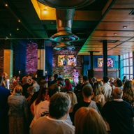 Crowd gathered inside a lively bar venue watching a live music performance, with stage lights and screens in the background.