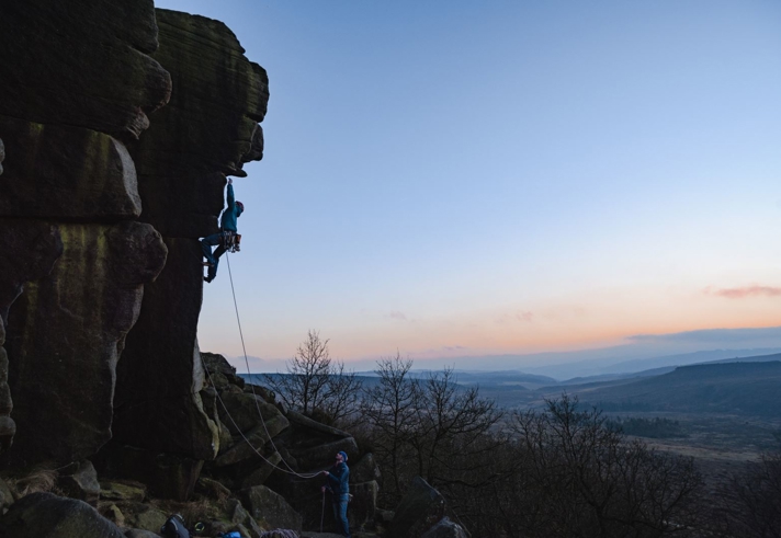 A person is rock climbing on a steep cliff face at dusk, secured by a rope held by another person standing below. The sky is clear with a gradient from blue to orange near the horizon, and the landscape in the background features rolling hills and sparse trees.