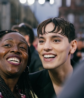 Two people standing close together in an outdoor setting, with a crowd and buildings visible in the background. The image shows bright stage lights overhead and blurred architectural details behind them, suggesting an event or gathering in an urban area. One person is wearing a dark outfit, while the other has braided hair and a patterned top.