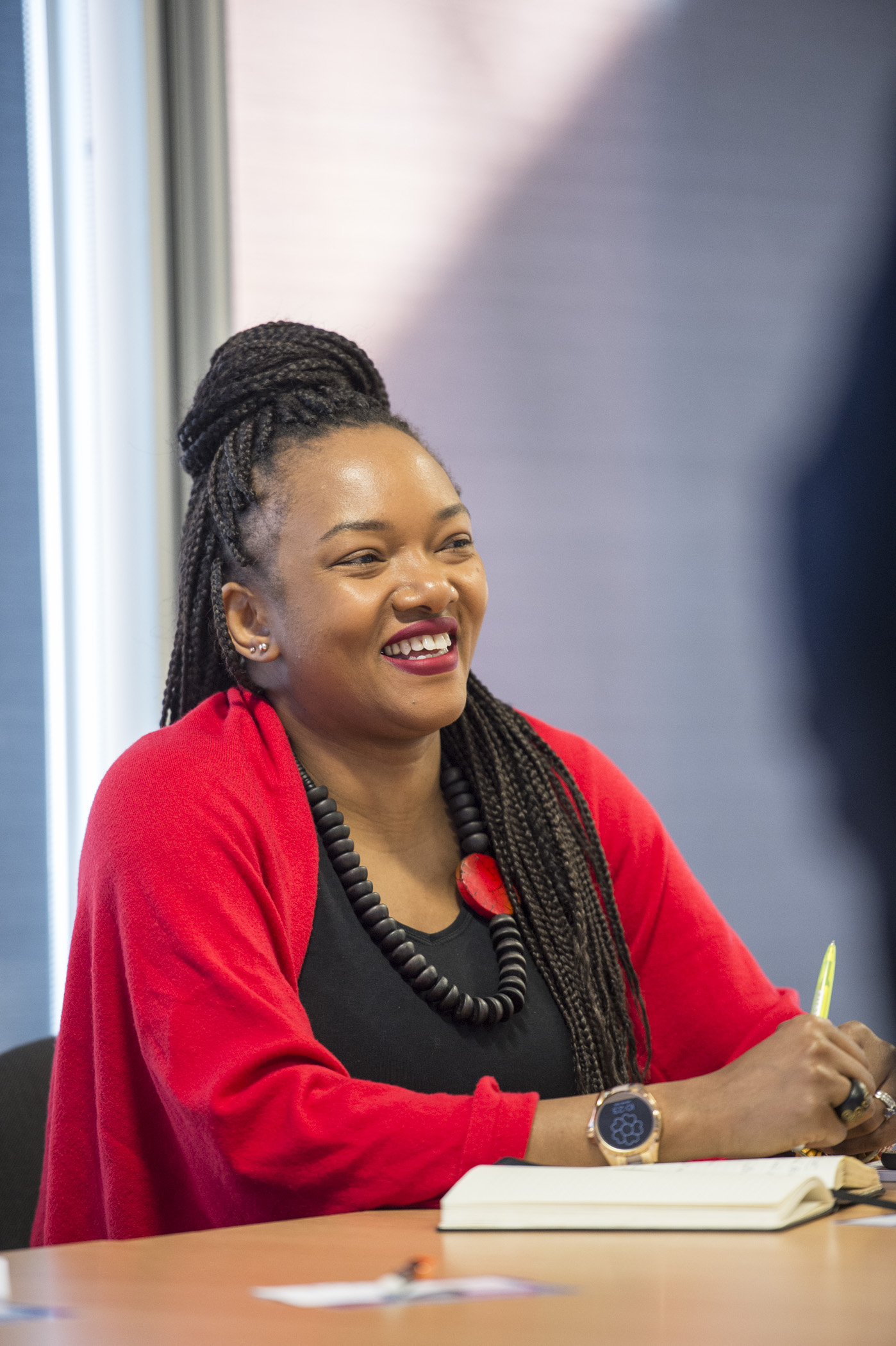 A middle-aged black woman in a red  cardigan and chunky jewellery sits in a meeting at a modern office. 