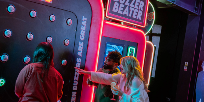 Three people playing an interactive arcade game called “Buzzer Beater” in a neon-lit venue. The game features a large vertical panel with multiple round buzzers and a digital score display reading “0550.” Bright signage on the machine includes phrases like “Green buzzers are great.” One person is actively playing, while another points at the panel, holding a drink. The setting is vibrant with colourful lights and a lively atmosphere.