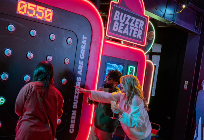 Three people playing an interactive arcade game called “Buzzer Beater” in a neon-lit venue. The game features a large vertical panel with multiple round buzzers and a digital score display reading “0550.” Bright signage on the machine includes phrases like “Green buzzers are great.” One person is actively playing, while another points at the panel, holding a drink. The setting is vibrant with colourful lights and a lively atmosphere.