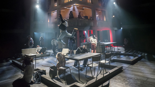 A dramatic stage performance featuring multiple actors in an industrial-style set. The foreground shows scattered tables and chairs with performers in dynamic poses, including one person leaping into the air. The background includes a two-level structure with additional actors visible on the upper floor, illuminated by theatrical lighting in shades of blue and red. The scene conveys high energy and movement.