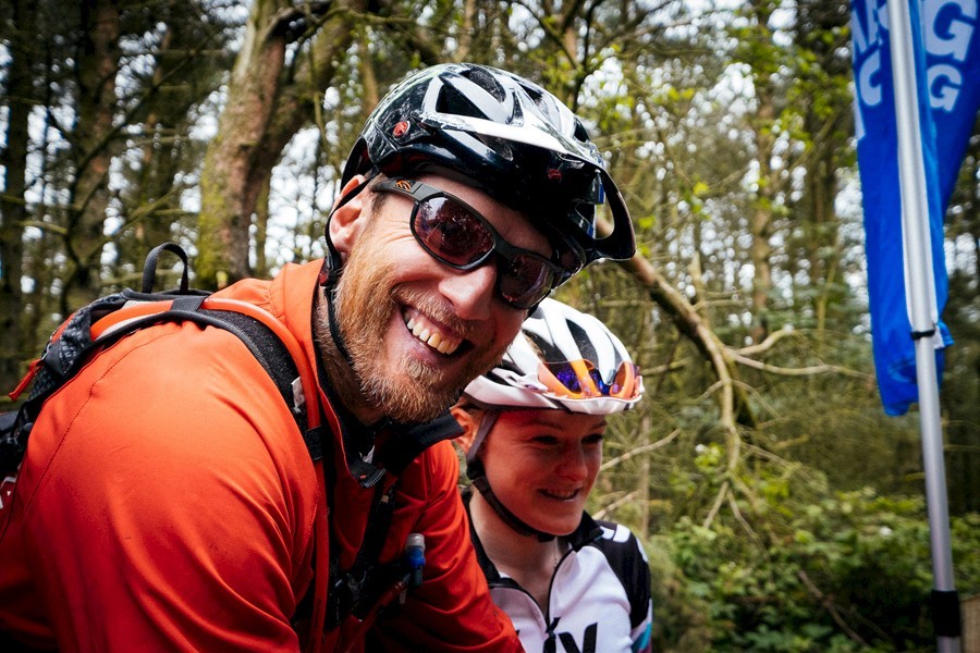 Two cyclists wearing helmets and outdoor gear, standing close together in a wooded area. One cyclist is dressed in a bright red jacket, and the other in a white cycling jersey with black and colored accents. A blue vertical banner is visible on the right side, and the background shows dense trees and greenery, suggesting a forest trail setting.