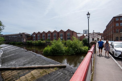 View from a red-railed bridge over a calm river with a small weir on the left. Two people walk along the bridge near parked cars. Across the river are historic red-brick mill buildings with pitched roofs and large windows, surrounded by greenery. A sign on one building reads ‘Kelham Island Museum.’ The sky is clear with light clouds.