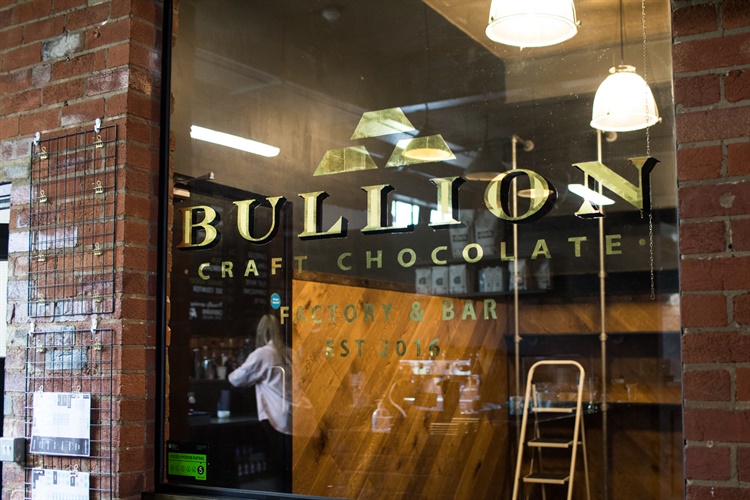 Storefront of Bullion Craft Chocolate, featuring a glass window with the gold Bullion logo and name. Inside, wooden shelves, a ladder, and hanging lights are visible. A person is seen through the window. The building has a red brick exterior.