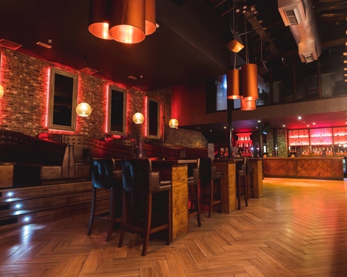 Interior shot of Crystal. Modern bar interior with warm lighting, exposed brick walls, and wooden herringbone flooring. The space features high tables with leather chairs, booth seating along the wall, and a long bar counter with illuminated shelves stocked with bottles. Copper pendant lights hang from the ceiling, adding to the stylish atmosphere.
