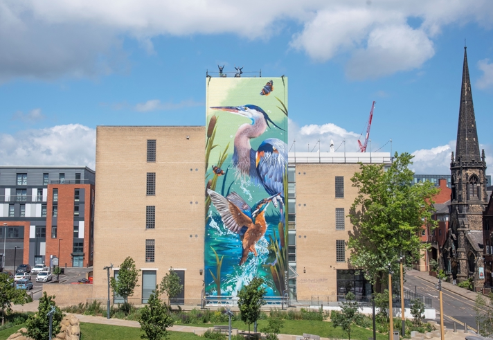 Large mural painted on the side of a tall brick building depicting vibrant wildlife, including a heron, a kingfisher, butterflies, and reeds in a water scene. The building is surrounded by green trees and a landscaped area, with modern apartments on the left and a historic church with a tall spire on the right under a partly cloudy blue sky.