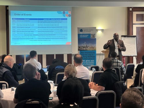 A speaker stands at the front of a conference room giving a presentation to an audience seated at tables. Behind the speaker is a large projection screen displaying an “Order of Events” schedule with times and topics, and a banner with the Sheffield Chamber logo and mission statement. A flip chart is positioned to the right of the speaker. The room has neutral walls, wooden panels, and bright overhead lighting.