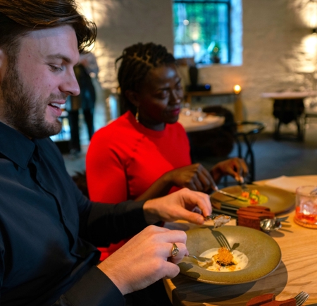 Two diners seated at a wooden table in a softly lit restaurant, eating a carefully plated dish with drinks beside them.