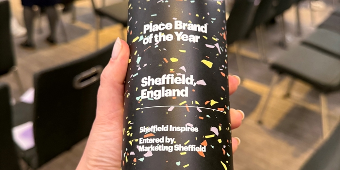 A hand holding a tall, cylindrical award with a colourful confetti‑style design. The text on the award reads “Place Brand of the Year – Sheffield, England – Sheffield Inspires – Entered by Marketing Sheffield – City Nation Place Awards 2025.” Rows of chairs and people mingling in the event space appear softly in the background.