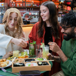 Three people seated at a round green table in a lively indoor food hall with a bar in the background. The table is filled with a wooden bento-style tray containing assorted sushi, nigiri, and pickled ginger, alongside plates of salad, bread, and spring rolls. Several tall glasses of colourful drinks with fruit and herb garnishes are also on the table. The setting is bright and vibrant, with shelves of bottles visible behind the bar.