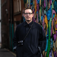 A man in a long, dark coat, with a camera slung over his shoulder, stands in front of some colourful street art.