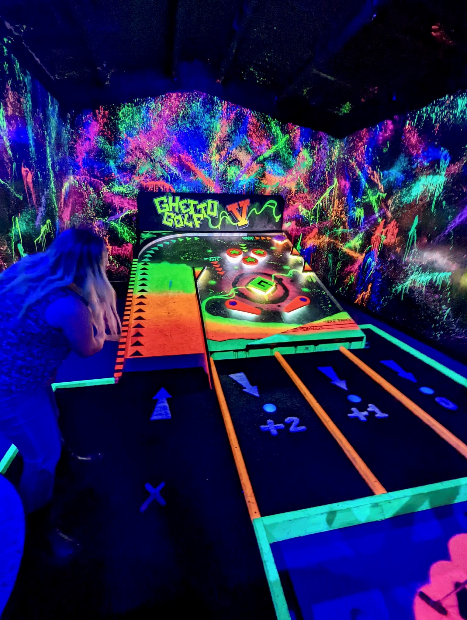 Woman taking a shot at on an indoor crazy golf course that looks like a giant pin ball machine and is illuminated with ultra violet lights 