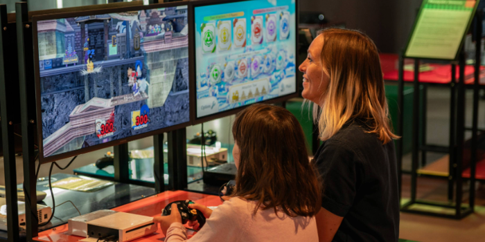 A person with a child sat next to them are playing a video game together. In the background are displays of video games from the history of the video game industry.