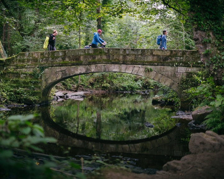 Three people walking across an old stone bridge in a lush forest setting. The bridge arches over a calm stream that reflects the greenery and the shape of the arch, creating a near-perfect circle in the water. The surrounding area is dense with trees, vines, and undergrowth, giving the scene a natural and tranquil atmosphere.