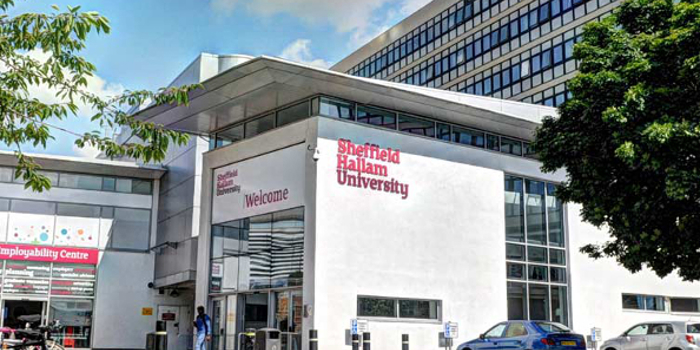 Entrance to Sheffield Hallam University featuring a modern building with large glass windows and white walls. The university's name is displayed in bold red letters. Bicycles are parked on the left, cars on the right, and a tree provides partial shade. People are walking near the entrance under a partly cloudy sky