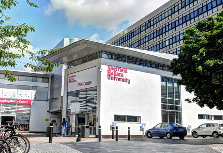 Entrance to Sheffield Hallam University featuring a modern building with large glass windows and white walls. The university's name is displayed in bold red letters. Bicycles are parked on the left, cars on the right, and a tree provides partial shade. People are walking near the entrance under a partly cloudy sky