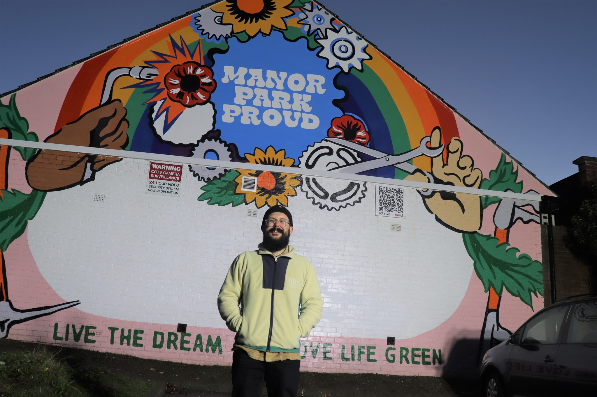 “Colorful mural on the side of a building featuring a rainbow, flowers, gears, and large hands holding tools, with the words ‘Manor Park Proud’ in bold white letters at the center. Below the mural, the text reads ‘Live the Dream, Live Life Green.’ A person stands in front of the artwork, and part of a car is visible on the right.”