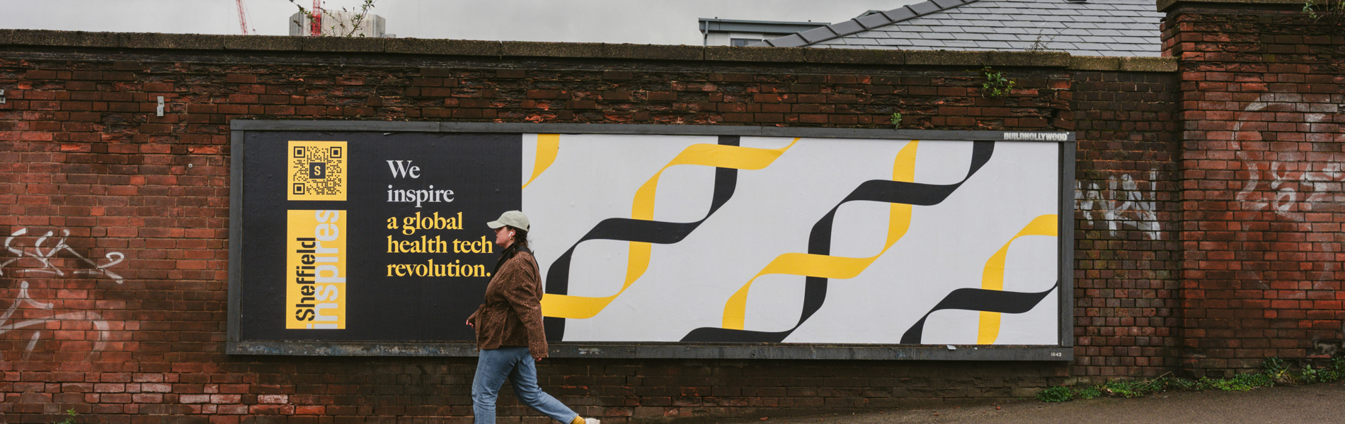 A person walks along a pavement in front of a large brick wall featuring a billboard. The billboard has a modern design with black and yellow geometric DNA-like patterns on a white background. On the left side, there is a black panel with yellow text that reads: “Sheffield” and “We inspire a global health tech revolution.” The sky is overcast, and there are construction cranes visible in the distance above the wall.
