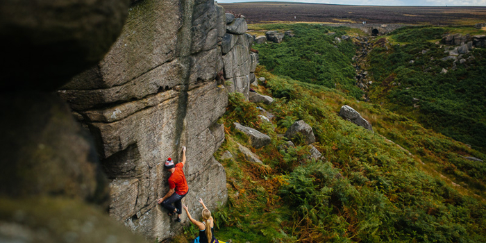 Two people are climbing up a rock face. They are using climbing gear and have a safety mat on the ground.