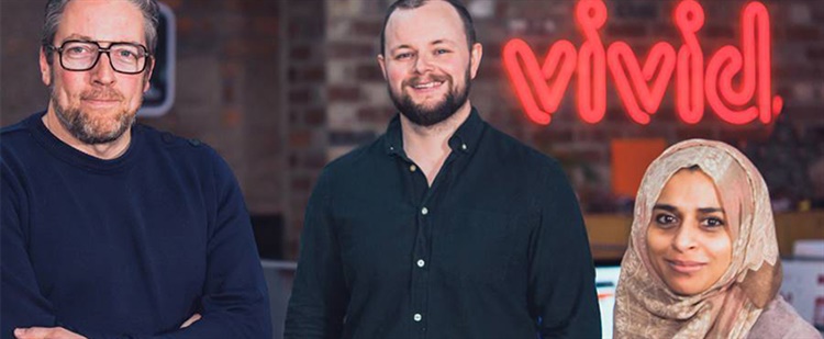 Two men and a woman standing in front od a red neon sign that reads 'Vivid'.