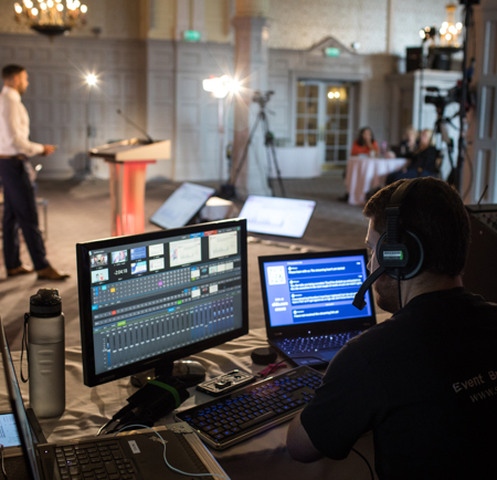 A live event production setup showing a technician operating multiple screens and audio equipment in the foreground, while a speaker stands at a podium in the background. The room is equipped with cameras, lighting, and monitors for broadcasting.