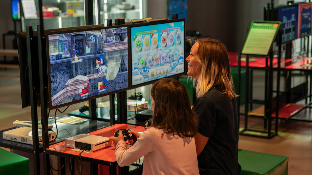 Two people sit at a gaming station inside the National Videogame Museum, playing a video game on two large screens. The setup includes controllers, a console, and bright red tables, with other gaming stations visible in the background.