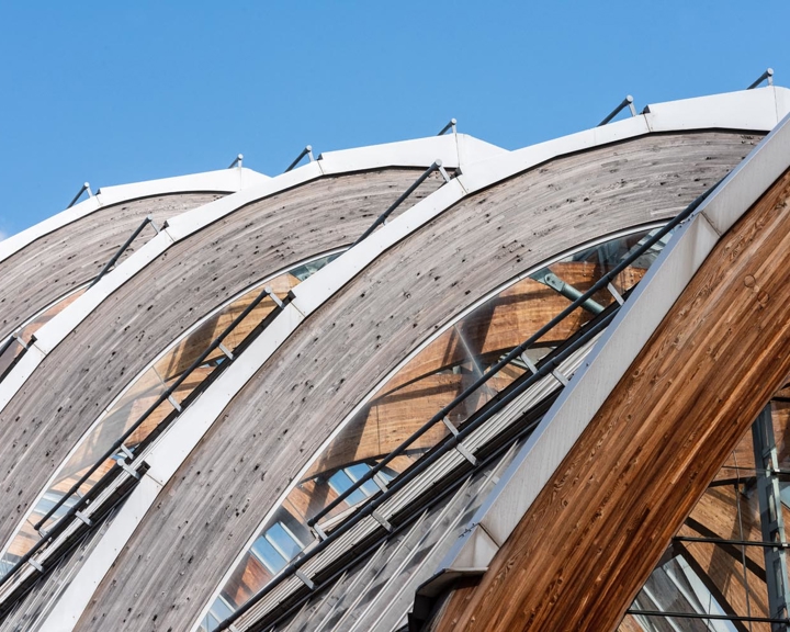 Close-up view of large curved wooden arches forming part of a modern architectural structure. The arches are supported by metal framework and glass panels, with reflections of surrounding buildings visible. The design features repeating geometric patterns against a clear blue sky.