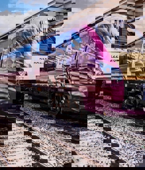 EMR Gold Award 222 - Modern maroon and cream-colored passenger train with the number 222104 parked at a station platform. The train has a sleek aerodynamic design with reflective windows, and the station features a covered roof structure with metal beams. Bright daylight and partly cloudy skies are visible in the background.