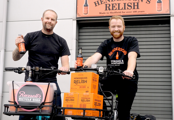 Two people standing with bicycles in front of a building featuring a sign that reads “Hendersons (Sheffield) Ltd Makers of Henderson’s Relish – Made in Sheffield for over 100 years.” Both bicycles have baskets filled with bright orange boxes labeled “Henderson’s Relish.” One person holds a bottle of Henderson’s Relish, and the other has a bottle in the basket. A smaller sign on one bike reads “Russell’s Bicycle Shed.” The background includes a grey roller shutter and the orange Henderson’s Relish branding.