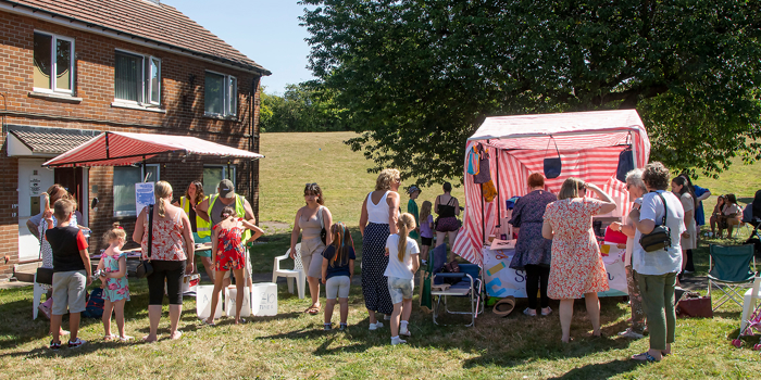 People gathered outdoors at a summer fair on a grassy area near a brick house. A red-and-white striped stall is set up under a large tree, with several individuals standing around it. Others are chatting or waiting nearby, and some children are present. Folding chairs and a small canopy are visible, suggesting a casual community event in warm weather.