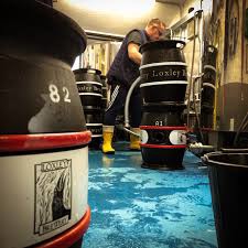 Person working in a brewery surrounded by black beer kegs labelled ‘Loxley Brewery,’ standing on a blue floor and wearing bright yellow boots.