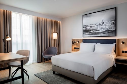 Modern hotel room with a large bed dressed in white linens, a wooden headboard with built-in lighting, and a black-and-white cityscape photo above the bed. The room includes a desk with a lamp and chair, an upholstered armchair, and floor-to-ceiling curtains covering a bright window.