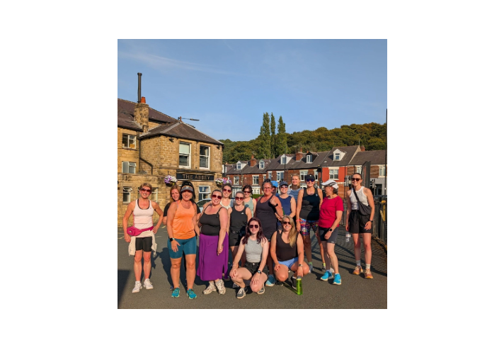 Group of 17 people in casual athletic wear standing on a street in front of a building named "The Abbey." The background includes houses and greenery on a hillside under sunny, clear weather.