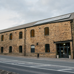 Exterior view of a large stone building with tall arched windows and a pitched roof fitted with solar panels. The structure has an industrial yet modern look, with a glass entrance door flanked by potted plants. It sits alongside a paved road with white markings, and the sky above is overcast. The building appears to be a converted mill or warehouse with contemporary features.