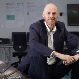A person wearing a dark navy suit with a white dress shirt, seated on a red cushioned examination table in a clinical setting. A stethoscope hangs around the neck, suggesting a medical professional. The background includes a light blue frosted glass panel and a section of white drawers on the right side.