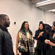 A small group of people standing indoors in conversation. The setting appears to be a modern room with white walls and exposed ceiling fixtures. One person is wearing a quilted black jacket, another has a large plaid scarf draped over their shoulders, and others are dressed in dark clothing. A white bench is visible along the wall in the background.