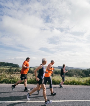 Four people jogging along a rural road on a bright day, with open countryside and green fields stretching into the distance under a partly cloudy sky.