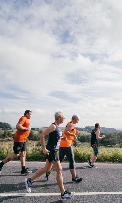 Four people jogging along a rural road on a bright day, with open countryside and green fields stretching into the distance under a partly cloudy sky.